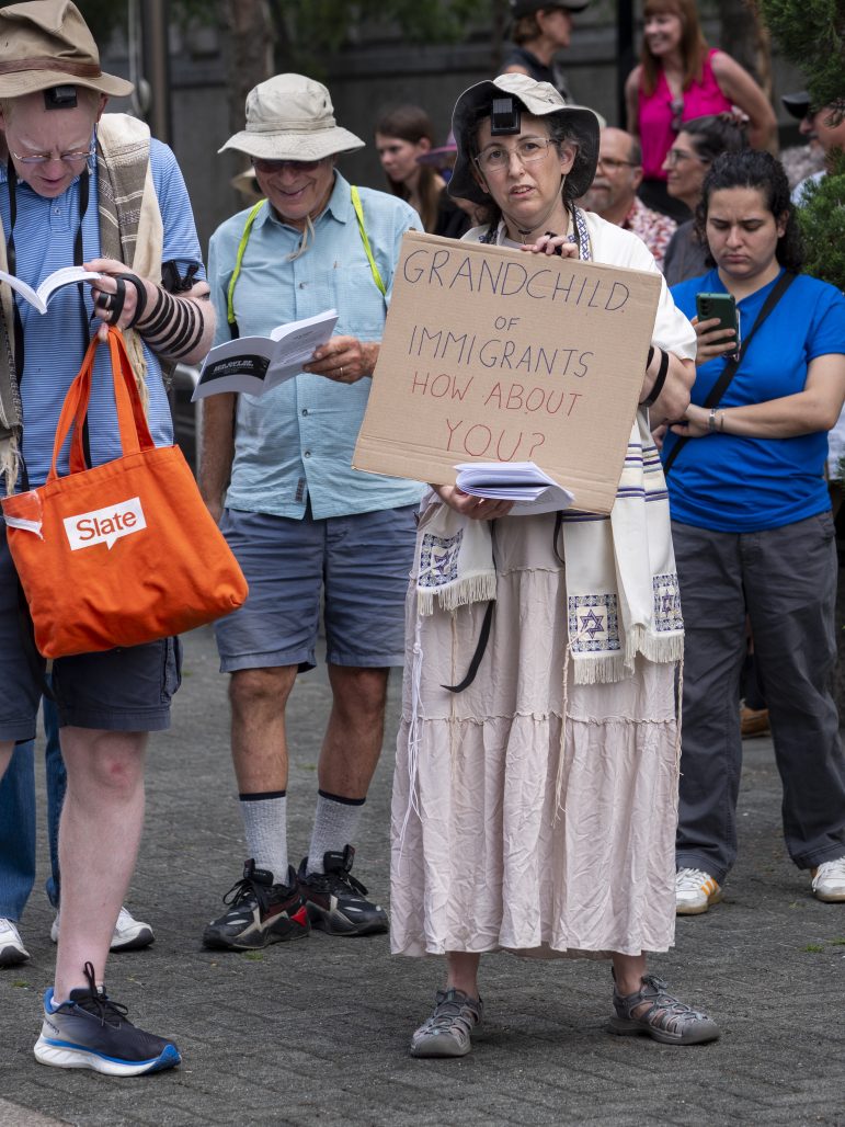 A person in a pale pink skirt wears a tallit and carries a sign that says "Grandchild of Immigrants. How about you?"